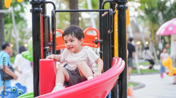 Fototapeta A young Asian boy playing happily on a colorful playground slide in a park during daylight. He shows joy, curiosity, and energy while exploring the play area. 
