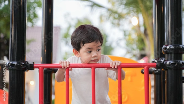 Fototapeta A young Asian boy playing happily on a colorful playground slide in a park during daylight. He shows joy, curiosity, and energy while exploring the play area. 