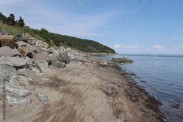 Fototapeta Vast beach and rocks in a bay and in summer. Charlevoix landscape in Quebec in Canada and riverside. Vacation and travel in Charlevoix. Shore and coastline with an horizon. Panorama and vast bay. 