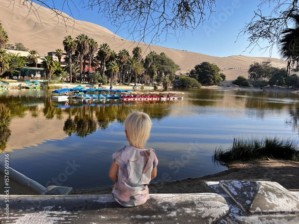 Fototapeta Little child or toddler sitting back to the camera and watching Huacachina lagoon surrounded by sand dunes desert, all reflected in the lake, Atacama desert, Ica, Peru, South America. 