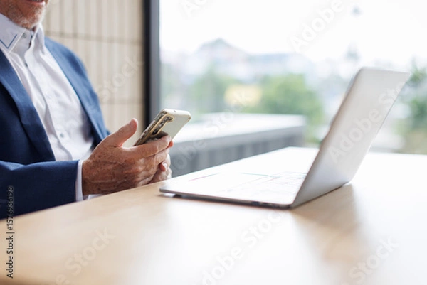 Obraz Portrait of a cheerful mid adult businessman using phone for investment plan chatting contact Investor searching for informationat at workplace office. connecting people concept