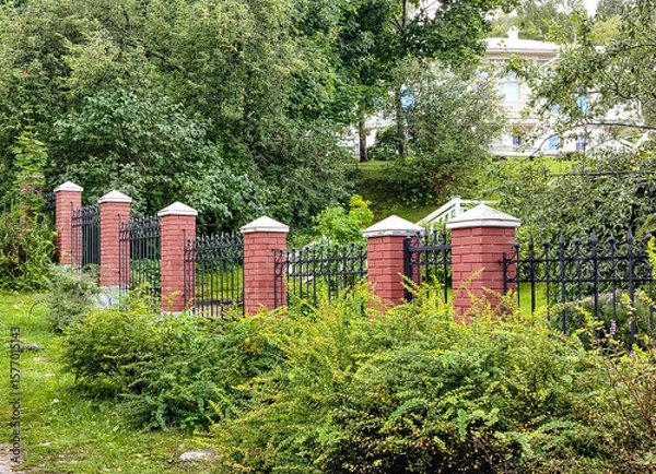 Fototapeta A wrought metal black fence with lattice and red brick posts separates the park from the lady on the hill behind the trees