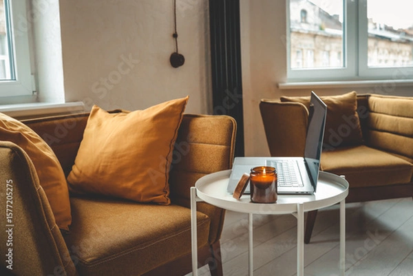 Fototapeta Cozy home interior with mustard armchair, burning candle in glass jar, and laptop on a small white table. Warm natural light and peaceful atmosphere