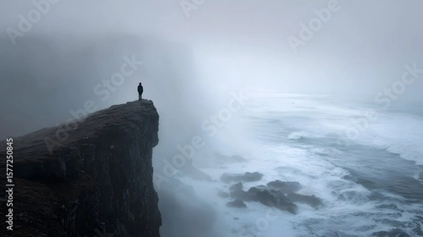 Fototapeta Person standing on a cliff edge overlooking the ocean under a moody, misty sky.