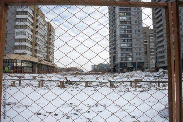 Fototapeta Snow covers a vacant construction site amid tall, modern buildings. A chain-link fence encloses the area, showcasing an incomplete urban project in a chilly environment.