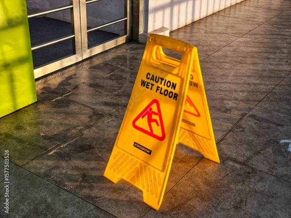 Obraz A bright yellow "Caution Wet Floor" sign stands on a shiny tiled surface, illuminated by warm sunlight. This safety sign is a common sight in public spaces, reminding people to be careful in slippery