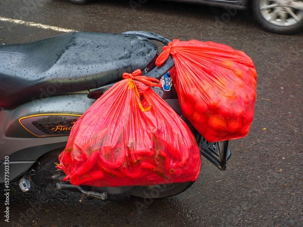 Obraz Two large red plastic bags filled with chili peppers and tomatoes are tied to the back of a motorcycle on a wet street. A real-life glimpse into traditional market transportation in urban settings.