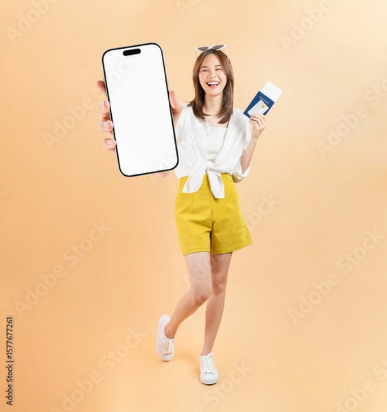 Fototapeta Happy smiling young Asian woman holding smartphone mockup of blank screen and passport with plane tickets and credit card on beige color background. Tourist travel trip journey concept.