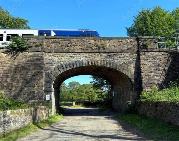 Fototapeta A Victorian stone bridge spans a dirt path beneath a passing train, framed by lush foliage and open sky, with sunlight sharpening the texture in Norwood Green, Halifax, UK.
