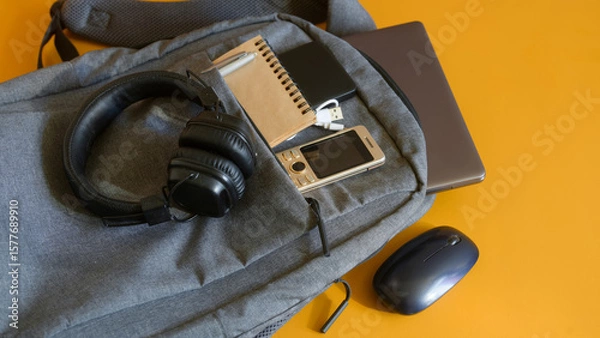 Obraz Modern study or travel setup featuring a roomy backpack with electronic gadgets—laptop, phone, powerbank, mouse, USB cable, notebook and cushioned headphones on yellow. High resolution