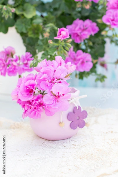 Fototapeta Pink geranium flowers arranged in a decorative purple vase with wooden flower embellishments, placed on a white doily, with additional geraniums visible in soft focus in the background,vertical