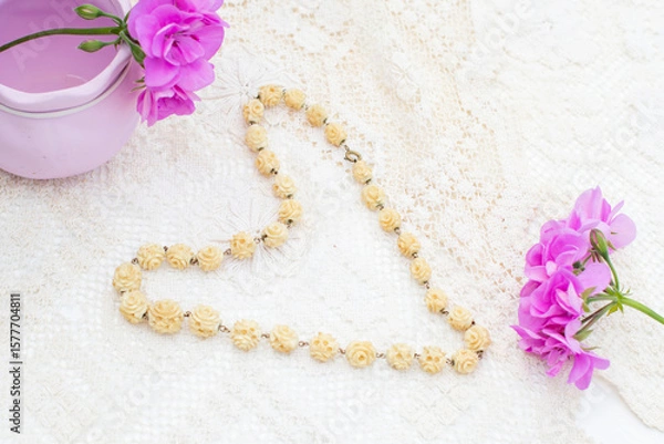 Fototapeta Vintage ivory necklace with rose-shaped beads lies on a white lace tablecloth in the shape of a heart, next to purple geranium flowers and a lilac vase, creating a delicate and romantic composition,