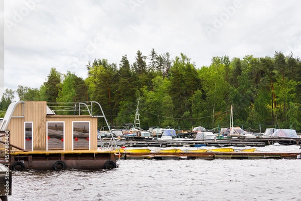 Fototapeta Floating wooden house and pier with boats lined up in order on lakeside shore waiting near green forest under cloudy sky