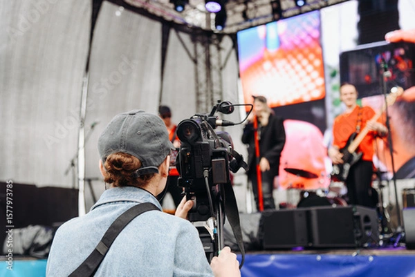 Fototapeta large black video camera on tripod, videographer working at music festival in city park, rock group performing on stage