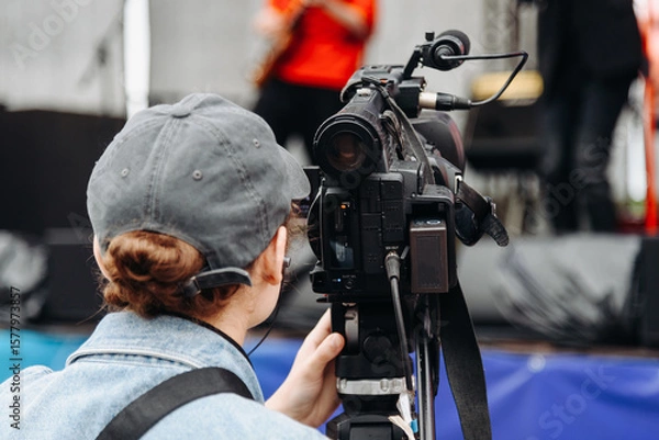 Fototapeta large black video camera on tripod, videographer working at music festival in city park, rock group performing on stage