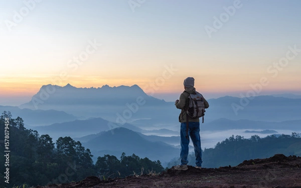 Fototapeta Male Hiker with backpack relaxing on top of rock mountain and enjoying sunset sky.