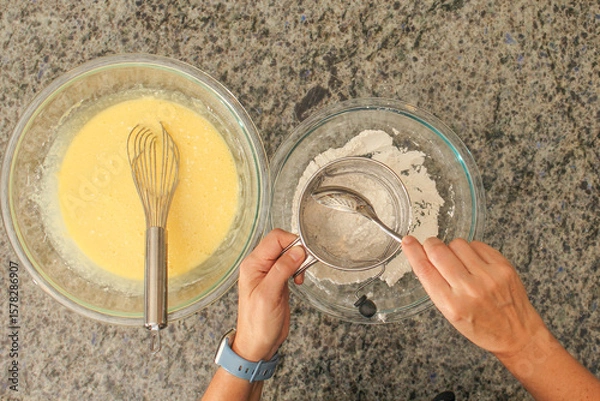 Fototapeta Overhead View of Hands Sifting Flour with Hand Crank Sifter for Baking