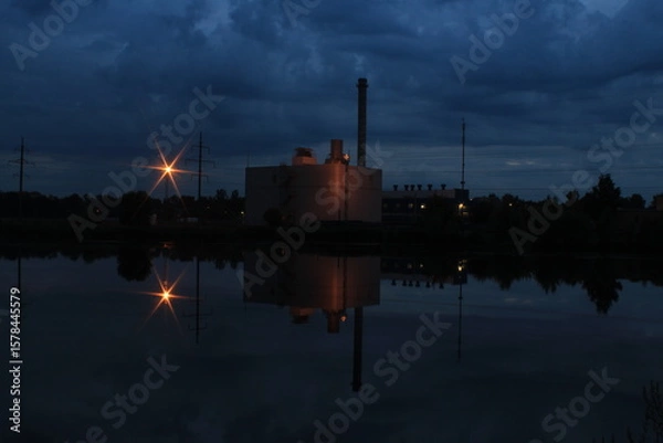 Fototapeta Industrial building with beautiful lighting brightly reflected in the water of the lake at dusk.