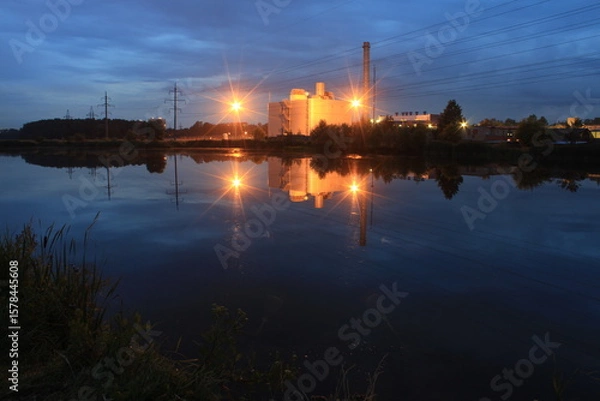 Fototapeta Industrial building with beautiful lighting brightly reflected in the water of the lake at dusk.