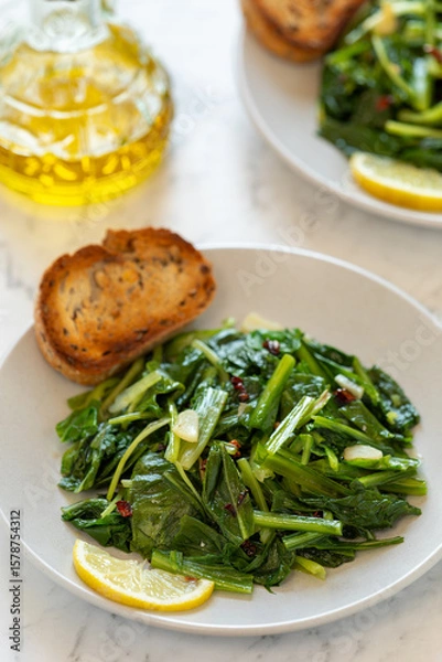 Fototapeta Roman-style sautéed chicory greens (Cicoria Ripassata alla Romana) served on a plate with toasted bread and a slice of lemon. A bottle of olive oil and a second plate are in the background.