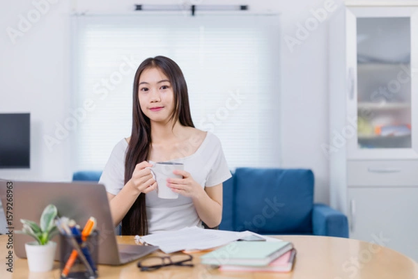 Fototapeta Beautiful Asian businesswoman enjoying coffee while working on a laptop at home, embodying the work-from-home lifestyle with a blend of comfort and modern technology