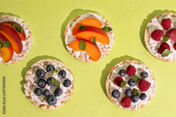 Obraz Rice cakes with different types garnish fruits, on green background. Healthy colorful food. Crispy puffed rice cakes with fruit and honey