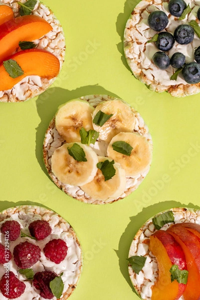 Obraz Rice cakes with different types garnish fruits, on green background. Healthy colorful food. Crispy puffed rice cakes with fruit and honey