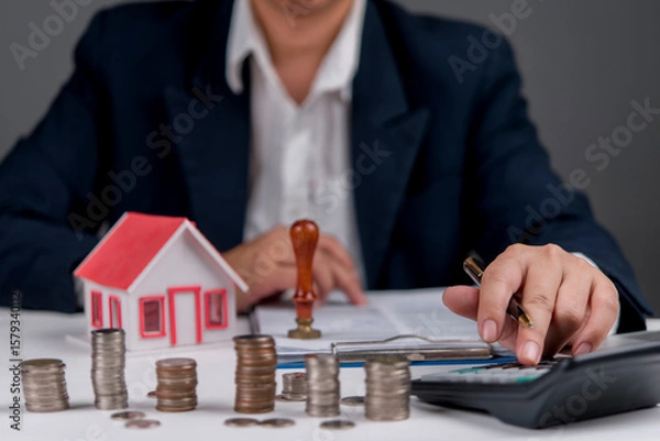 Fototapeta Businessman calculating real estate investment, with house model, coins, and calculator, symbolizing property finance, mortgage planning, home loan, asset growth, financial advisor.