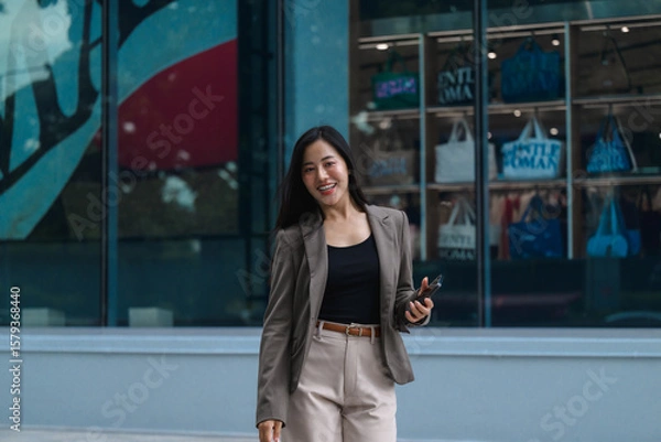 Fototapeta Smiling businesswoman walking and using smartphone in front of modern building