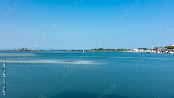 Fototapeta Peaceful lagoon view in Grado, Italy, with calm waters, a stony shoreline, and distant boats under a bright summer sky.