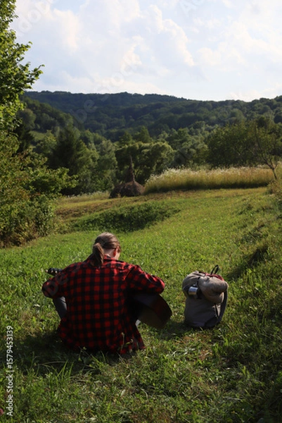 Obraz A young guy in a red shirt and jeans with a guitar travels alone through green mountains and nature.