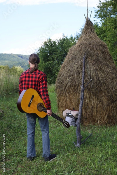 Obraz A young guy in a red shirt and jeans with a guitar travels alone through green mountains and nature.