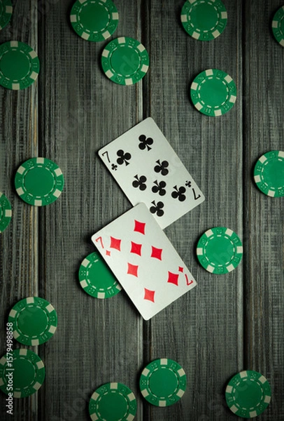 Fototapeta Two playing cards featuring the number two are placed amidst green poker chips on a wooden surface, showcasing an engaging card game atmosphere at night