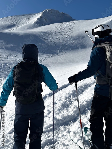 Obraz Two skiers admire a pristine mountain slope carved with fresh tracks under a clear blue sky. The thrill of untouched powder and alpine freedom.
