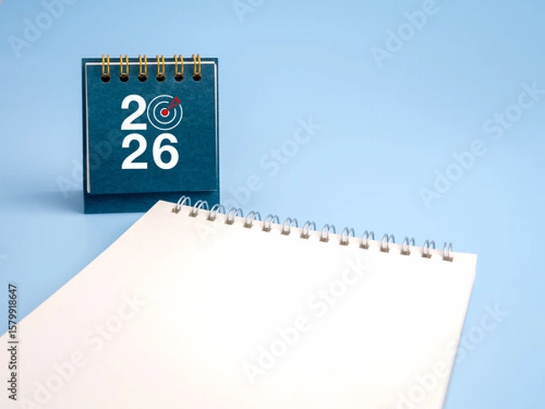 Obraz Happy new year 2026. 2026 numbers with target dart icon on blue desk calendar cover, standing near white blank notepad, empty space note page for memo. Business goal, resolutions and success concept.