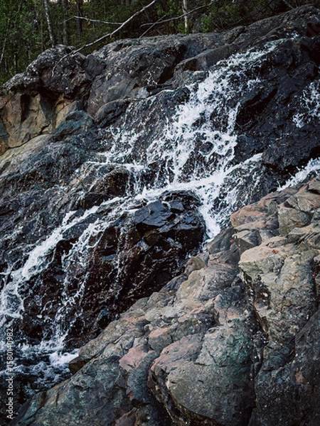 Fototapeta A waterfall is flowing down a rocky cliff. The water is clear and the rocks are grey. The scene is peaceful and serene