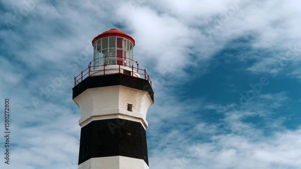Fototapeta Close up view of historic lighthouse with a red roof and black-and-white tower stands tall under a bright blue sky with scattered clouds, guiding ships along the Cape Recife coast in South Africa.