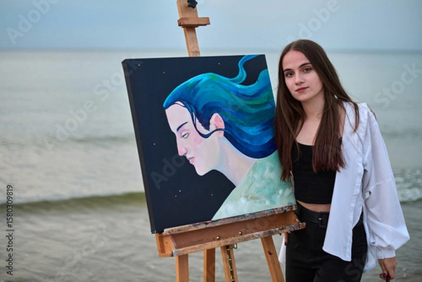 Fototapeta Young female painter stands by ocean, showcasing her vibrant portrait on canvas. Serene background complements her artistic expression, evoking tranquility and creativity