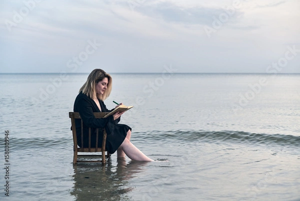 Fototapeta Young woman sits on chair in shallow water at sunset, writing in notebook. Serene ocean setting evokes feelings of peace and introspection, blending creativity and nature