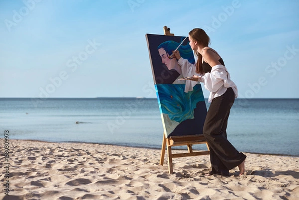 Fototapeta Young woman artist paints vibrant portrait on sandy beach, capturing tranquil ambiance of seaside. Her focused expression and calm sea evoke creativity and peace