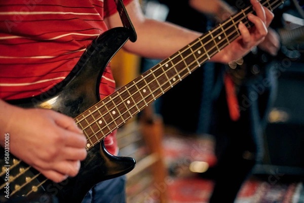 Fototapeta Focused view of musician hand playing bass guitar during performance, highlighting strings and frets in dim setting, musical rehearsal for new band member