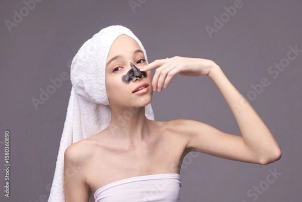 Fototapeta Young caucasian female applying charcoal face mask with towel on head for skincare routine.