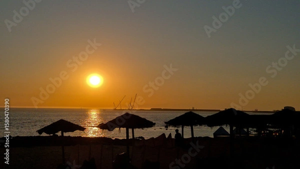 Fototapeta Sunset at Homem do Leme beach in Porto, Portugal, on July 8, 2025, showing silhouetted beach umbrellas and cranes against a golden sky, capturing a peaceful holiday evening by the Atlantic Ocean.