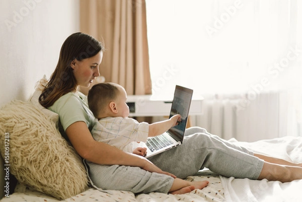 Fototapeta Portrait of happy young mother with little baby using laptop at home. Together, smiling mom browsing internet on computer while spending time with her little son.