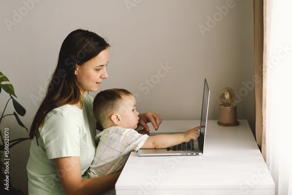 Fototapeta Portrait of happy young mother with little baby using laptop at home. Together, smiling mom browsing internet on computer while spending time with her little son.