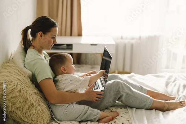 Fototapeta Portrait of happy young mother with little baby using laptop at home. Together, smiling mom browsing internet on computer while spending time with her little son.