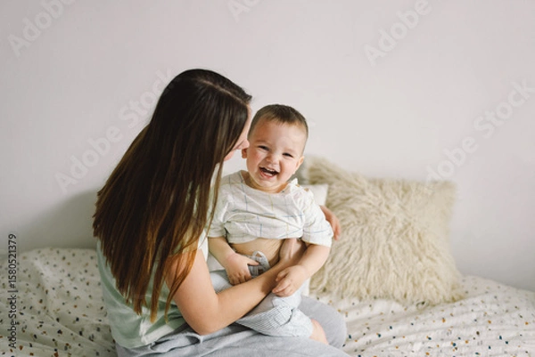Fototapeta Home portrait of a baby boy with mother on the bed relaxing and playing in the bed at the weekend together, lazy morning, warm and cozy scene. Mom holding and kissing her child. Mother day concept