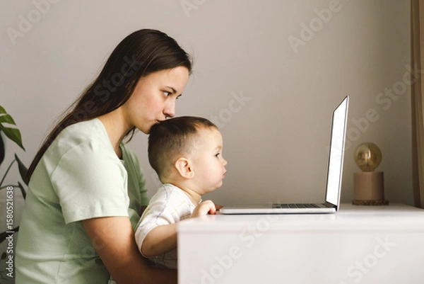 Fototapeta Portrait of happy young mother with little baby using laptop at home. Together, smiling mom browsing internet on computer while spending time with her little son.