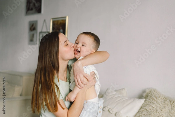 Fototapeta Home portrait of a baby boy with mother on the bed relaxing and playing in the bed at the weekend together, lazy morning, warm and cozy scene. Mom holding and kissing her child. Mother day concept