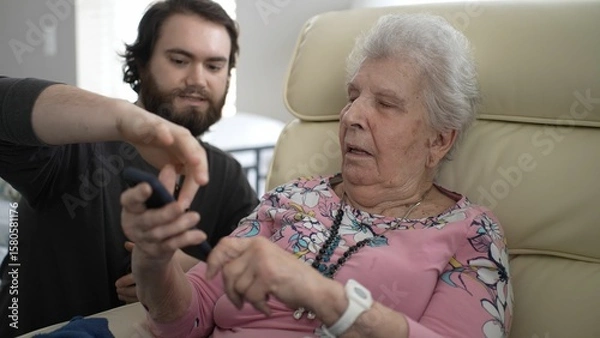 Fototapeta In a cozy living room, a grandson is guiding his grandmother to share photos on her phone, creating happy memories together. Their expressions show joy and connection.